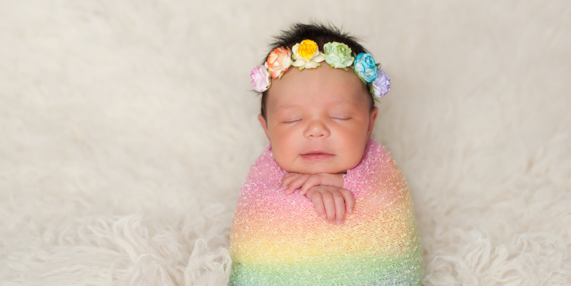 Baby laying on blanket wrapped in rainbow swaddle and wearing a flower headband