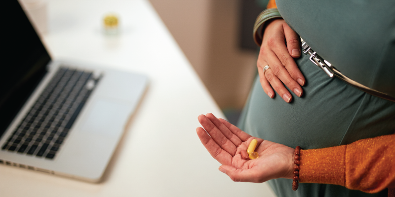 Pregnant woman standing by laptop and holding vitamins